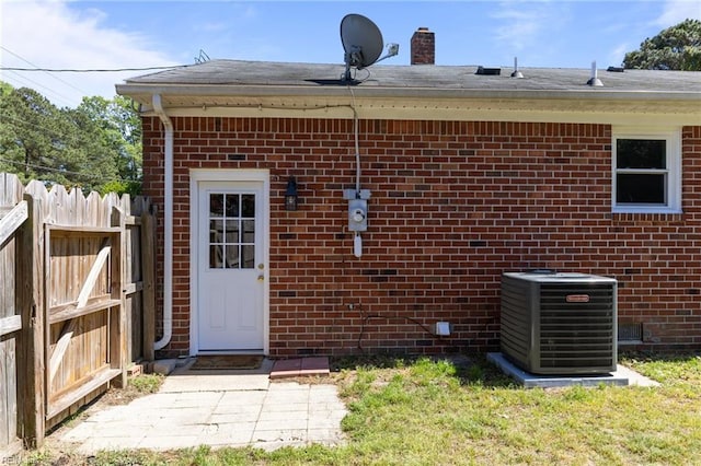 doorway to property featuring fence, a shingled roof, central AC, brick siding, and a chimney