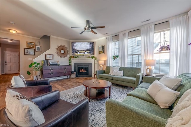 living room featuring a ceiling fan, visible vents, wood finished floors, a glass covered fireplace, and ornamental molding