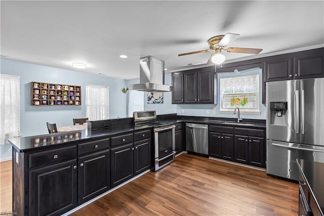 kitchen featuring a peninsula, dark countertops, island exhaust hood, a sink, and appliances with stainless steel finishes