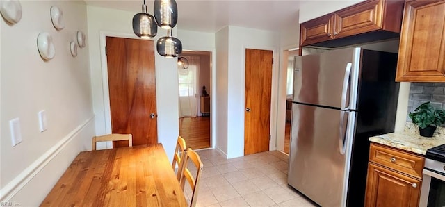 kitchen featuring freestanding refrigerator, light stone countertops, light tile patterned floors, brown cabinets, and backsplash