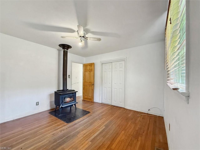 unfurnished living room featuring a ceiling fan, baseboards, wood finished floors, and a wood stove