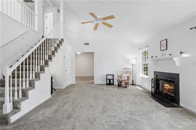 carpeted living room with a ceiling fan, visible vents, a fireplace with flush hearth, high vaulted ceiling, and stairs