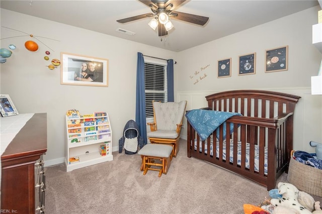 bedroom with a crib, carpet, a wainscoted wall, a ceiling fan, and visible vents
