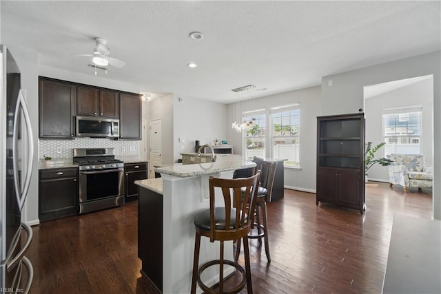 kitchen with decorative backsplash, appliances with stainless steel finishes, dark wood-style floors, a breakfast bar, and an island with sink