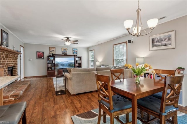 dining area with crown molding, a brick fireplace, ceiling fan with notable chandelier, wood finished floors, and visible vents