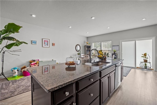 kitchen with light wood-style flooring, dark stone countertops, a sink, a center island with sink, and stainless steel dishwasher