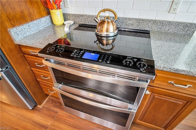 kitchen view of brown cabinets, tasteful backsplash, range with two ovens, light stone counters, and light wood-style flooring