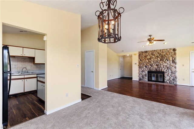 unfurnished living room with a stone fireplace, dark colored carpet, a sink, ceiling fan with notable chandelier, and dark wood-style floors
