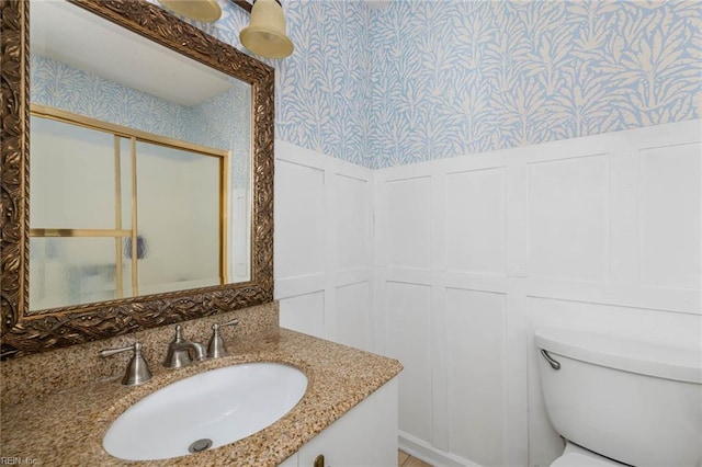bathroom with wallpapered walls, a decorative wall, a wainscoted wall, toilet, and vanity
