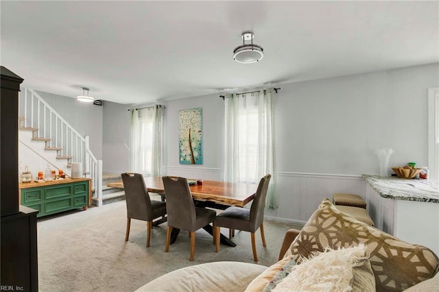 dining room featuring light colored carpet, stairs, a wainscoted wall, and a healthy amount of sunlight