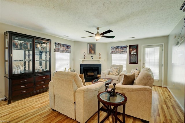 living area with ceiling fan, a fireplace, light wood-style floors, a textured ceiling, and baseboards