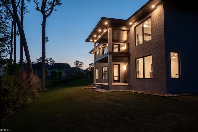 back of house at dusk with a yard, fence, brick siding, and a balcony