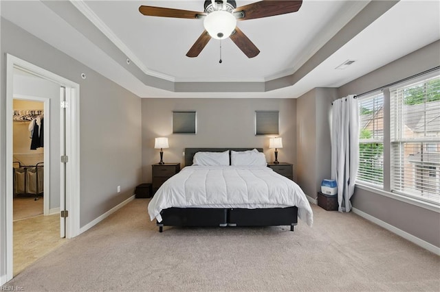 bedroom featuring light colored carpet, a tray ceiling, ornamental molding, baseboards, and a walk in closet