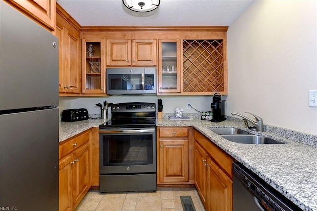 kitchen with a sink, brown cabinetry, light stone counters, glass insert cabinets, and appliances with stainless steel finishes