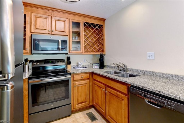 kitchen featuring glass insert cabinets, a sink, brown cabinetry, light stone countertops, and appliances with stainless steel finishes