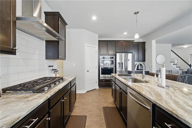 kitchen with dark brown cabinets, a sink, stainless steel appliances, light stone countertops, and wall chimney range hood