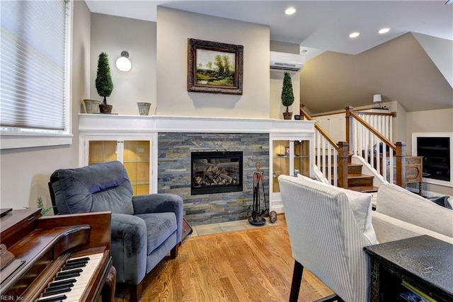 living area with a wall mounted AC, wood finished floors, a stone fireplace, recessed lighting, and stairway