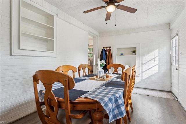dining area featuring a ceiling fan, built in shelves, brick wall, and wood finished floors