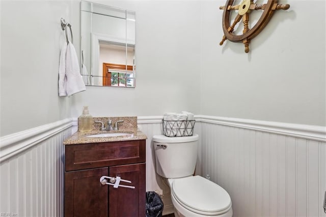 half bathroom with vanity, toilet, a decorative wall, and wainscoting