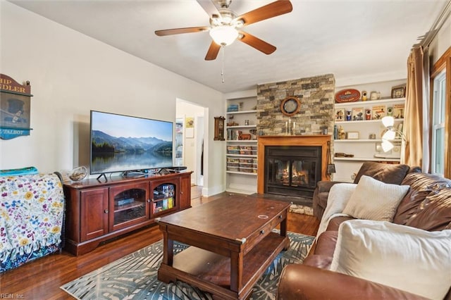 living area with a stone fireplace, dark wood-type flooring, a ceiling fan, and built in shelves