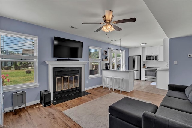 living room featuring light wood-style flooring, a ceiling fan, a fireplace with flush hearth, visible vents, and baseboards