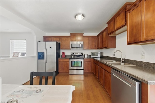 kitchen with appliances with stainless steel finishes, a sink, brown cabinets, dark countertops, and light wood-type flooring