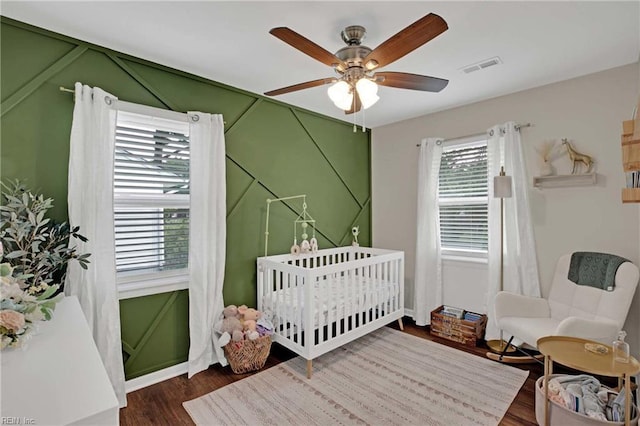 bedroom with dark wood-style flooring, a crib, and ceiling fan
