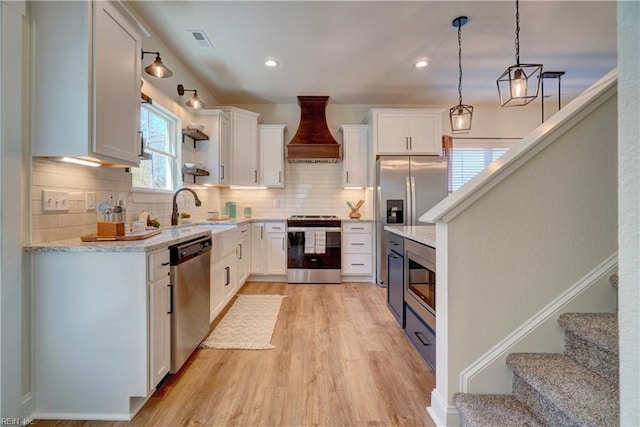 kitchen with appliances with stainless steel finishes, a sink, premium range hood, white cabinets, and light wood-type flooring