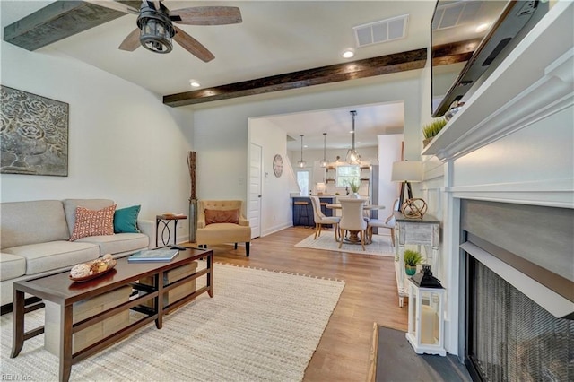 living area with ceiling fan, visible vents, a fireplace with flush hearth, wood finished floors, and beam ceiling