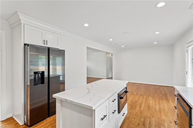 kitchen featuring stainless steel fridge, black oven, white cabinets, light wood-type flooring, and light stone countertops