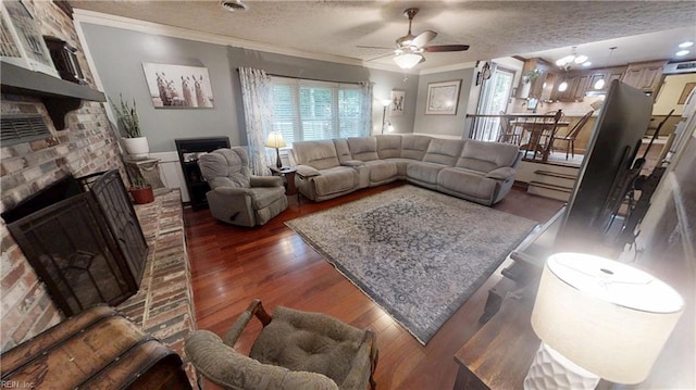 living room with a ceiling fan, a brick fireplace, a textured ceiling, dark wood finished floors, and crown molding