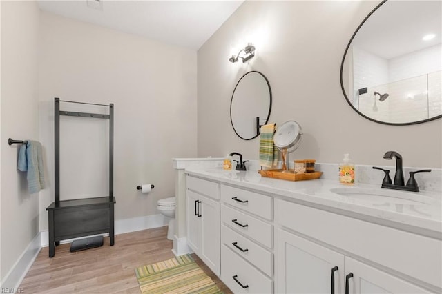 bathroom featuring wood finished floors, toilet, double vanity, a tile shower, and baseboards