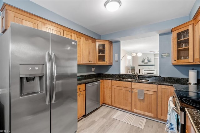 kitchen with stainless steel appliances, a sink, light wood finished floors, dark stone counters, and glass insert cabinets