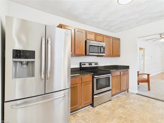 kitchen featuring appliances with stainless steel finishes, brown cabinetry, dark stone counters, light flooring, and baseboards