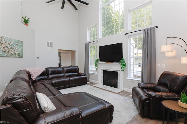 living room featuring a towering ceiling, wood finished floors, a ceiling fan, a fireplace with raised hearth, and baseboards