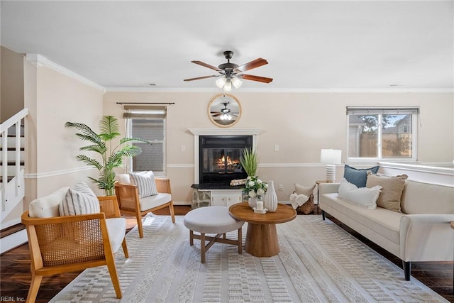 living area with a glass covered fireplace, ceiling fan, ornamental molding, wood finished floors, and baseboards
