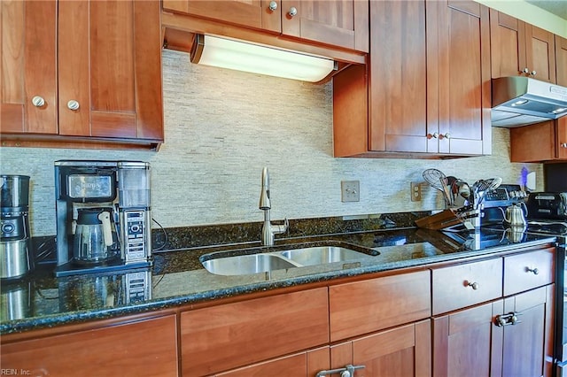 kitchen featuring a sink, under cabinet range hood, tasteful backsplash, brown cabinets, and dark stone counters