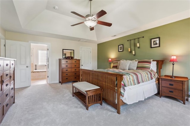 bedroom with a tray ceiling, light colored carpet, ensuite bathroom, and a ceiling fan