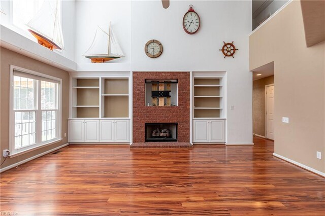 unfurnished living room with a towering ceiling, built in shelves, wood finished floors, baseboards, and a brick fireplace