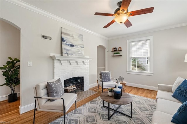 living area featuring arched walkways, light wood-type flooring, crown molding, a ceiling fan, and a brick fireplace
