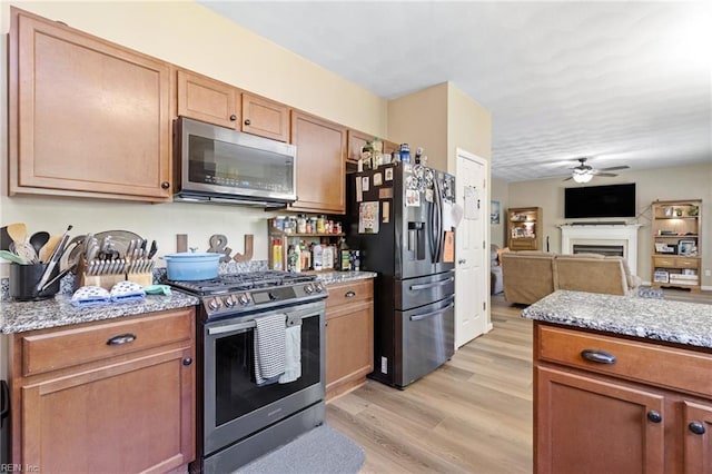 kitchen with appliances with stainless steel finishes, a fireplace, light wood-style flooring, ceiling fan, and light stone counters
