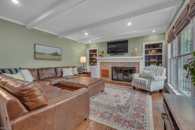 living room with wood finished floors, beam ceiling, a brick fireplace, built in shelves, and crown molding