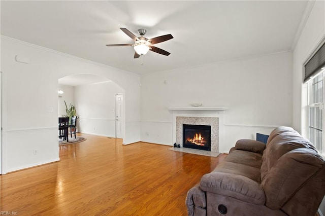 living room featuring arched walkways, a ceiling fan, crown molding, light wood-style floors, and a fireplace with flush hearth