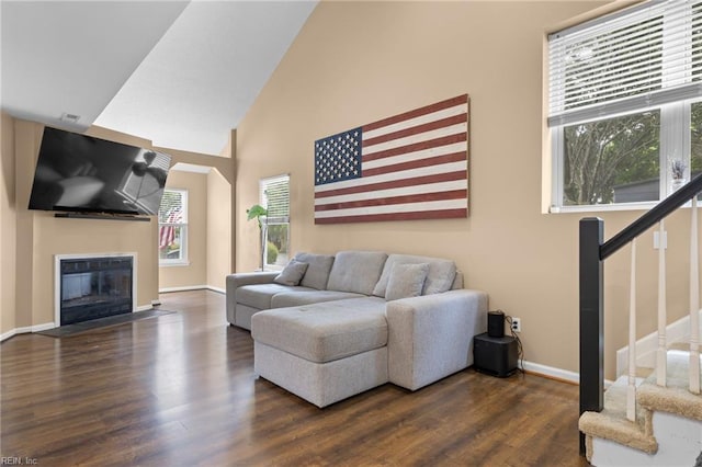 living area featuring stairs, dark wood-style flooring, baseboards, a glass covered fireplace, and high vaulted ceiling