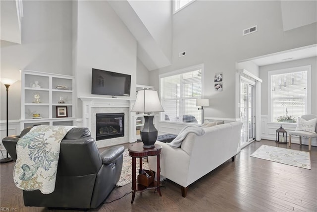 living room with a towering ceiling, healthy amount of natural light, wood finished floors, a glass covered fireplace, and a wainscoted wall