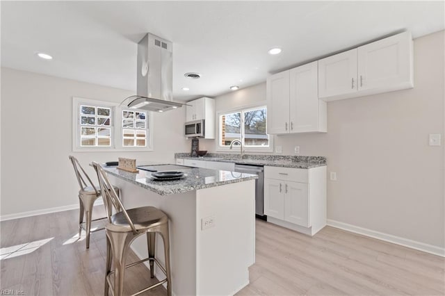 kitchen featuring island range hood, appliances with stainless steel finishes, a kitchen breakfast bar, light stone countertops, and light wood-style flooring