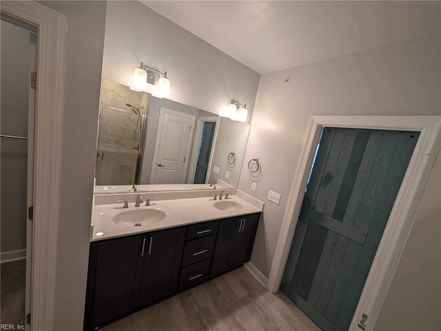 full bathroom with wood finished floors, double vanity, a tile shower, and baseboards