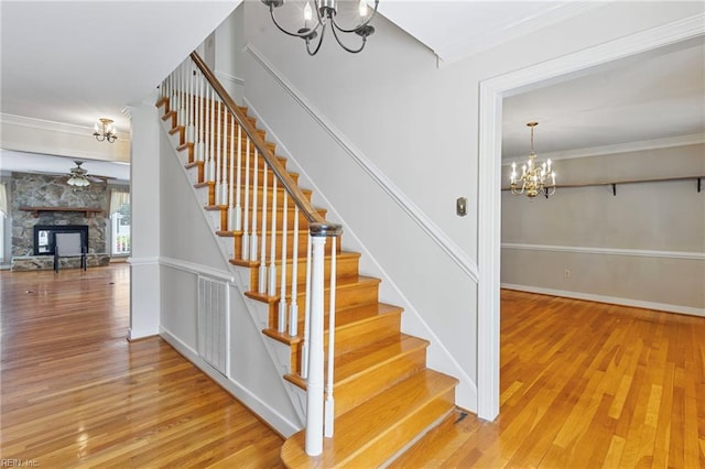 stairs with a chandelier, a stone fireplace, wood finished floors, crown molding, and a ceiling fan