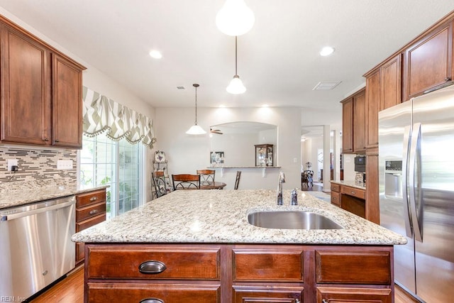 kitchen featuring stainless steel appliances, a sink, a kitchen island with sink, tasteful backsplash, and light stone counters