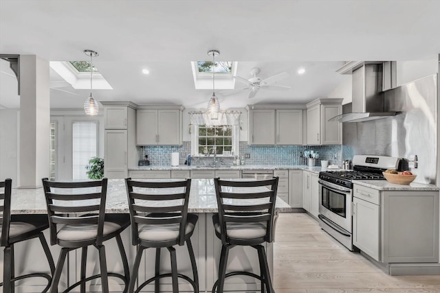 kitchen featuring gray cabinetry, light stone counters, a skylight, wall chimney range hood, and stainless steel gas range